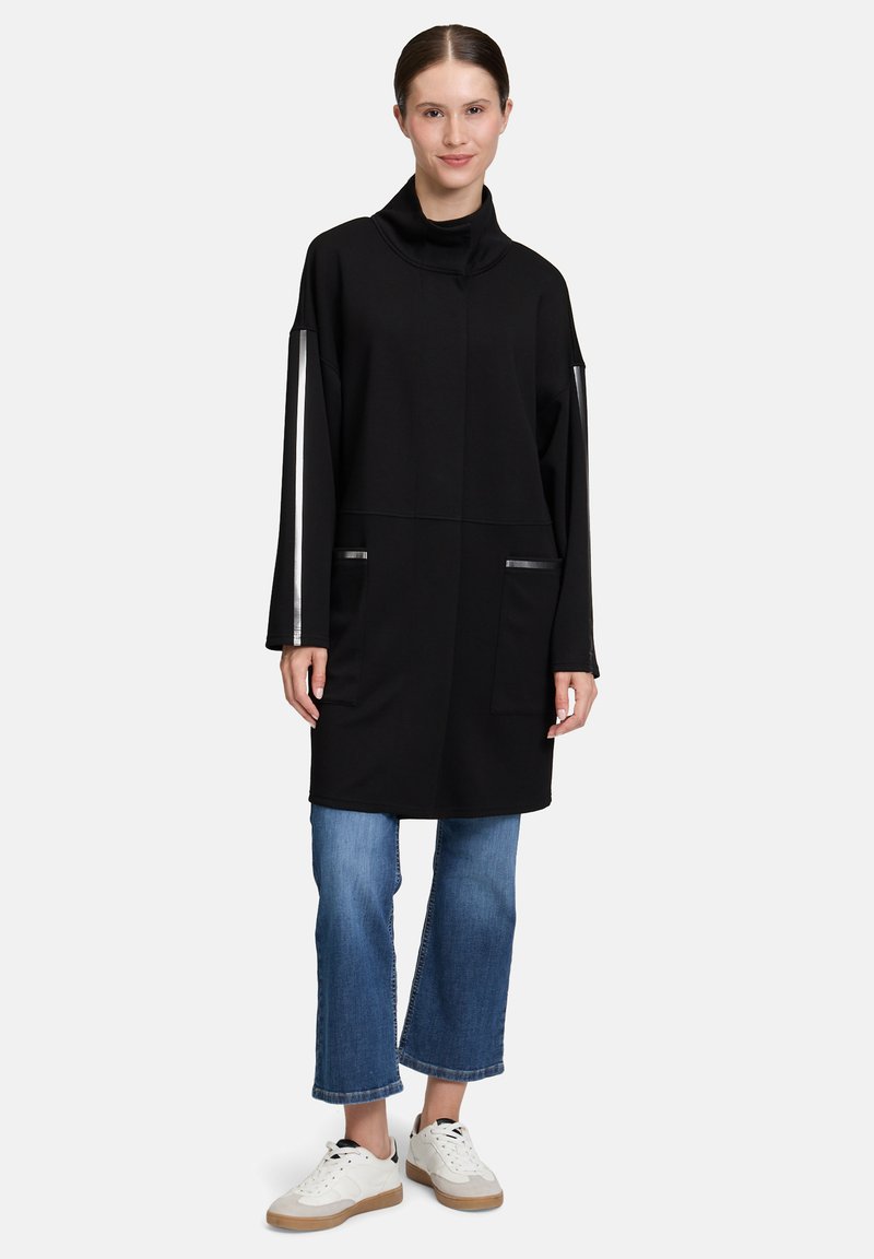 Frau in einer schwarzen Langarm-Tunika mit weißen Streifen an den Ärmeln, blauen, taillierten Jeans und weißen Sneakers, die vor einem schlichten Hintergrund steht.