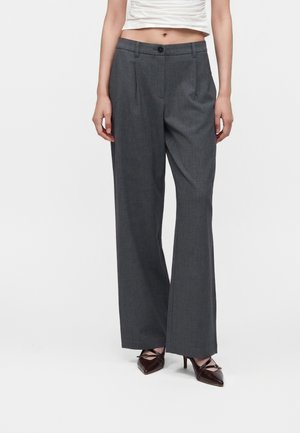 Femme portant un pantalon large taille haute gris foncé, un top court blanc et des talons pointus foncés, debout sur fond blanc.