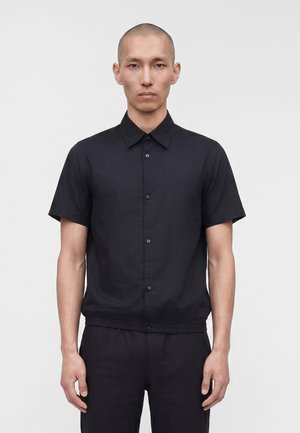 Lühikeste juustega mees, kes kannab musta lühikeste varrukatega nööbitavat särki ja musti pükse, seisab ühevärvilise valge tausta ees.