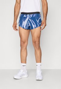 Blaue gemusterte Sportshorts mit schwarzem Bund, getragen mit weißen Socken und Sneakers. Hat ein welliges Design und eine figurbetonte Passform.
