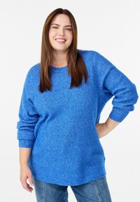 Kobieta z długimi, brązowymi włosami, uśmiechająca się, w niebieskim swetrze z dzianiny i niebieskich dżinsach, stojąca z jedną ręką na biodrze na tle białego tła.