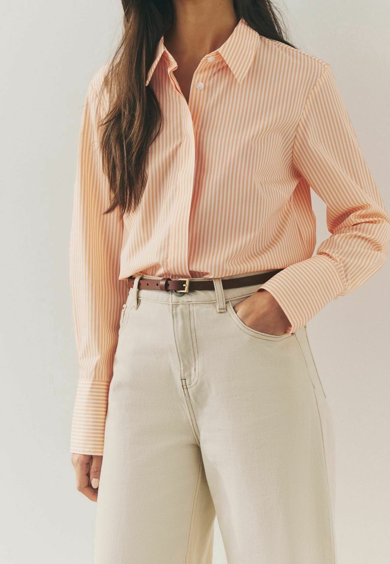 Chemise boutonnée à rayures pêche et blanc, dotée d'un col classique et de manches longues, associée à un pantalon taille haute beige clair.