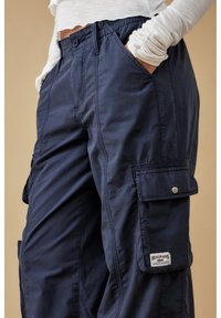 Person i marinblå cargobyxor med fickor och en vit långärmad topp, händerna i fickorna, beige neutral bakgrund.