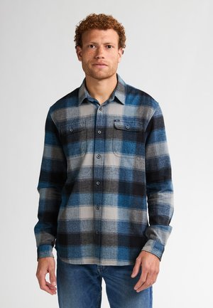 Camisa de franela para hombre en un patrón de cuadros azul y gris, con dos bolsillos en el pecho, cierre de botones al frente y mangas largas. Textura suave y corte holgado.