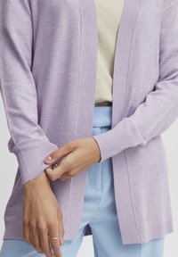 Světle fialový pletený cardigan s žebrovanými manžetami a otevřeným předním designem, nošený přes béžový top a světle modré kalhoty.