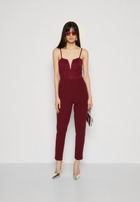 Burgunderfarget jumpsuit med blondebodice, slanke stropper og skreddersydde bukser. Tilbehør inkluderer en liten håndveske og metalliske høye hæler.