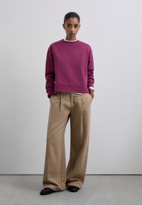 Una mujer se encuentra de pie contra una pared simple vistiendo un sudadera púrpura, pantalones beige de pierna ancha, zapatos negros y una pulsera de plata con las manos en los bolsillos.