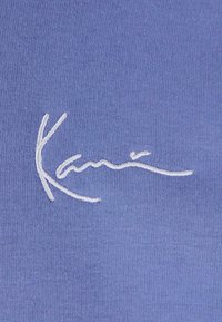 Broderie cursive blanche sur tissu bleu montrant une signature ou un détail de logo stylisé.