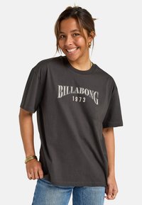 Camiseta de algodón gris oscuro con "BILLABONG 1973" en letras blancas. Corte holgado, mangas cortas, con un escote redondo y textura suave.