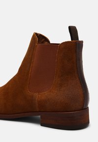Bottines Chelsea en daim brun avec des panneaux latéraux élastiques, une languette, et un talon en bois. Présentent une texture lisse et un design simple.