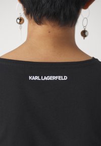 Musta t-paita, jossa valkoisella brodeerattu "KARL LAGERFELD" -teksti selkäpuolella. Silkkinen puuvillamateriaali, pyöreä kaula-aukko ja rento istuvuus.