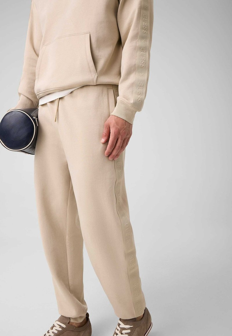 Personne portant un survêtement beige Guess et des baskets marron, tenant un petit sac rond noir, debout devant un fond uni gris clair.