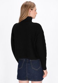 Zwarte gebreide trui met hoge kraag en textuurpatroon, gecombineerd met een donkere denimrok met achterzakken en contrasterende stiksels.