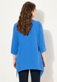 Blaue Tunika mit dreiviertel Ärmeln und abgerundetem Saum. Hergestellt aus leichtem Stoff mit glatter Textur, über schwarzen Hosen getragen.