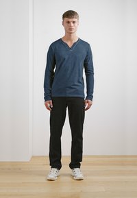 Marineblaues Langarm-V-Ausschnitt-Shirt kombiniert mit schwarzen Hosen. Das Modell trägt weiße Sneakers. Einfaches Design, weicher Stoff und taillierte Form.