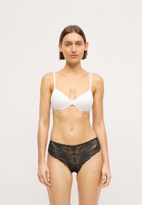 Woman with short brown hair wearing a white bra and black lace panties, standing against a plain light background, facing forward.