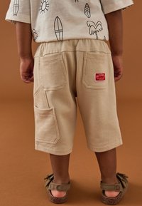 Smėlinės spalvos medvilniniai šortai su elastinga liemene, turintys dvi galines kišenes, šonines kišenes ir raudoną audinio etiketę. Dėvimi su rudomis sandalais.