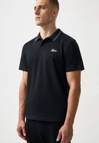 Svart polo skjorta i mjukt tyg, med en knappslå med två knappar, kontrasterande vita ränder på kragen och broderad logotyp på bröstet.