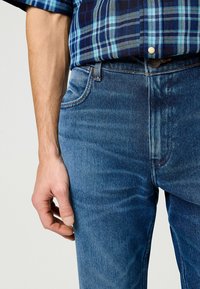 Donkerblauw geruite shirt, laagjes over blauwe denim jeans. De jeans hebben een klassiek vijf-pocket ontwerp met subtiele vervaging en stikdetails.