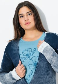 Marineblauer Cardigan mit Farbverlauf zu hellgrauen Bündchen, mit tiefem V-Ausschnitt, getragen über einem türkisfarbenen T-Shirt mit Blumenmuster. Glatte Textur.