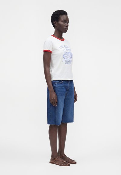 Γυναίκα φοράει λευκό T-shirt με κόκκινο περίγραμμα και γραφικό πίτσας, τζιν σορτς μέχρι το γόνατο και καφέ επίπεδα σανδάλια, στέκεται προφίλ.