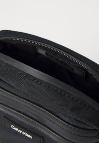 Borsa nera Calvin Klein con chiusura con zip, tessuto liscio e un'etichetta logo discreta all'interno. L'interno è compatto e foderato.