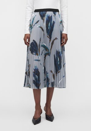 Jupe midi plissée avec un imprimé floral bleu et marron, portée avec un top blanc à manches longues et des talons noirs à bouts pointus.
