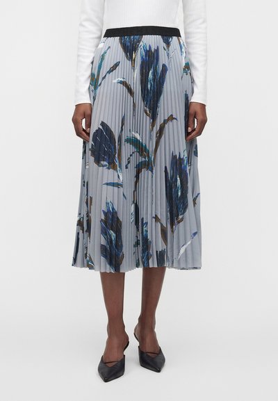 Jupe midi plissée avec un imprimé floral bleu et marron, portée avec un top blanc à manches longues et des talons noirs à bouts pointus.