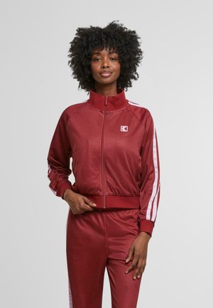 Giacca sportiva bordeaux con colletto alto, chiusura zip e strisce bianche ai lati. Realizzata in un tessuto testurizzato con un design traspirante.