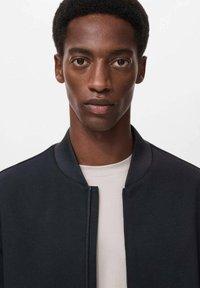 Chaqueta bomber negra con una textura suave y cuello redondo, llevada sobre una camisa de color beige claro. El diseño es minimalista y moderno.