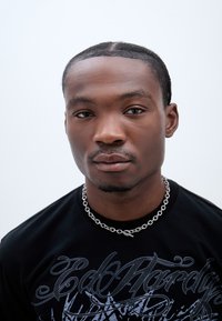 Fekete, rövid ujjú póló szürke grafikai mintával és ezüstlánc nyaklánccal. Közelkép egy arcról, egyenes hajjal, apró fonatokkal.