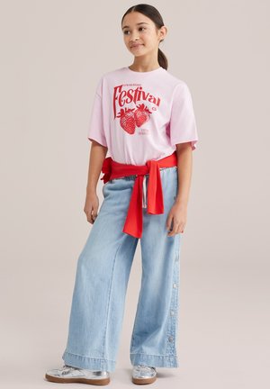 Mlada djevojka u ružičastoj majici s natpisom "Strawberry Festival", širokim svijetloplavim trapericama sa bočnim gumbima, srebrnim cipelama i crvenim džemperom vezanim oko struka.