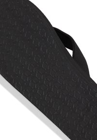 Musta flip-flopin pohja, joka esittelee rakenteellista pintaa geometrisilla kuvioilla. Hihna ja pohja ovat valmistettu kumimateriaalista, mikä korostaa kestävyyttä.