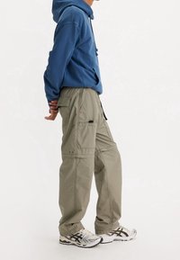 Cargo broeken in licht olijfgroen, met meerdere zakken, verstelbare taille en subtiele stiksels. Gecombineerd met een blauwe hoodie en hardloopschoenen.