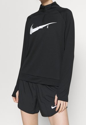 Osoba ubrana w czarny, długi rękaw Nike pulower z białym logo oraz czarne spodenki sportowe z białym znakiem Nike.