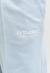 Tissu bleu clair avec le texte blanc "JJ Studios" et une petite accroche imprimée, montrant la texture et les détails des coutures du vêtement.