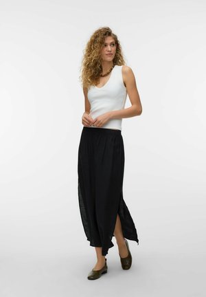 Kobieta z kręconymi włosami, ubrana w biały bezrękawnik, długą czarną spódnicę z bocznymi rozcięciami i oliwkowo-zielone baleriny, stojąca na jednolitym tle.