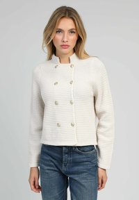 Veste blanche texturée avec un col montant, un design croisé et des boutons décoratifs en or. Associée à un jean bleu.