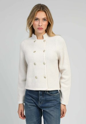Veste blanche texturée avec un col montant, un design croisé et des boutons décoratifs en or. Associée à un jean bleu.