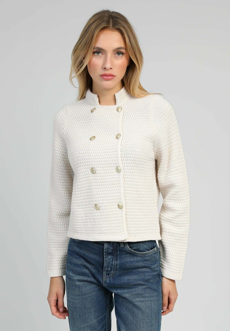 Veste blanche texturée avec un col montant, un design croisé et des boutons décoratifs en or. Associée à un jean bleu.