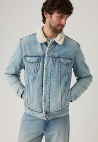 Lichtblauwe denimjas met een crèmekleurige nepbontkraag, knoopsluiting aan de voorkant, twee borstzakken en zichtbare stiksels.