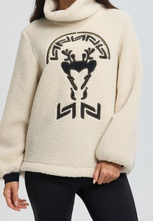 Kremowy polarowy sweter z czarnym geometrycznym wzorem renifera, noszony przez osobę w czarnych spodniach, stojącą z jedną ręką zrelaksowaną, a drugą chwytającą za dół.