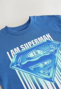Sininen puuvillainen t-paita pyöreällä kaula-aukolla, jossa on teksti "MINÄ OLEN SUPERMAN" ja suuri sininen Superman-logo graafisena, sekä pystysuorat vaaleansiniset raidat.