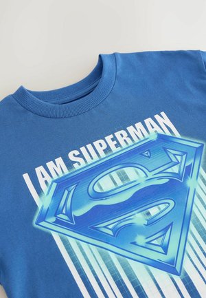 Mėlynas medvilninis marškinėlis su apvaliu kaklu, ant kurio yra užrašas "AŠ ESU SUPERMENAS" ir didelis mėlynas Supermeno logotipo grafikas, su vertikaliomis šviesiai mėlynomis juostelėmis.