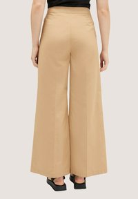 Kalhoty s širokými nohavicemi v světle hnědém materiálu, s vysokým pasem a texturovaným povrchem, nošené s černými platformovými sandály, pohled zezadu.