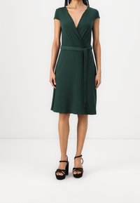 Abito verde avvolgente con scollatura a V profonda, maniche corte e cintura regolabile in vita. Tessuto testurizzato che arriva sopra il ginocchio, abbinato a tacchi neri.