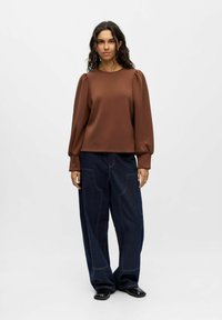 Braune Bluse mit langen Ärmeln und Puffärmeln, kombiniert mit lockeren, dunkelblauen Weitbeinjeans, die sichtbare Nähte und seitliche Taschen haben.