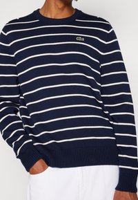 Granatowy sweter dzianinowy z białymi poziomymi paskami, ściągaczami przy mankietach i hemie oraz małym zielonym logo Lacoste na piersi.