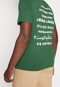 Zelené bavlněné tričko s potiskem vícerojazyčného textu na zádech. Krátké rukávy a klasický výstřih ve tvaru crew.
