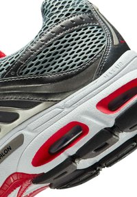 Zapato deportivo con parte superior de malla en gris y negro, con detalles en rojo, amortiguación de aire y una entresuela blanca texturizada.